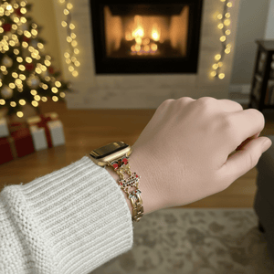 Hand wearing a gold watch with a Christmas-themed bracelet in a cozy room with a fireplace and tree.